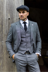 Homem de terno cinza e boné encostado em uma parede de madeira.