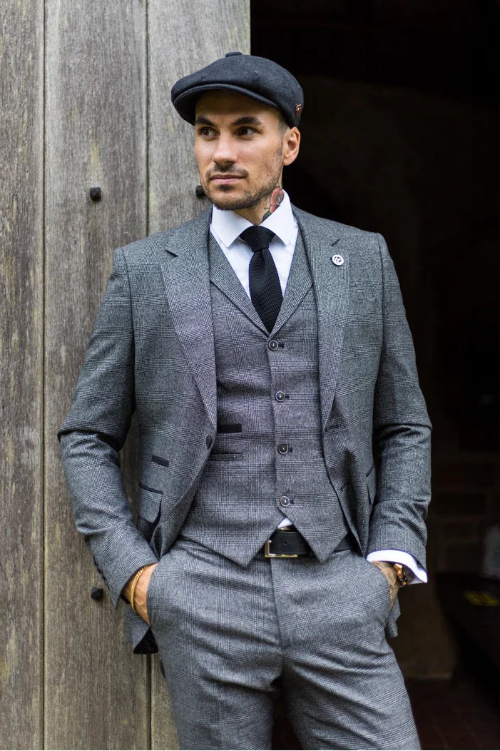 Homem de terno cinza e boné encostado em uma parede de madeira.