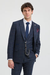 Homem vestindo um terno xadrez azul com camisa branca e gravata azul sobre um fundo cinza claro.