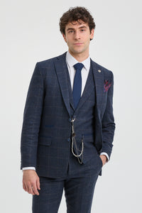 Homem vestindo um terno xadrez azul com camisa branca e gravata azul sobre um fundo cinza claro.