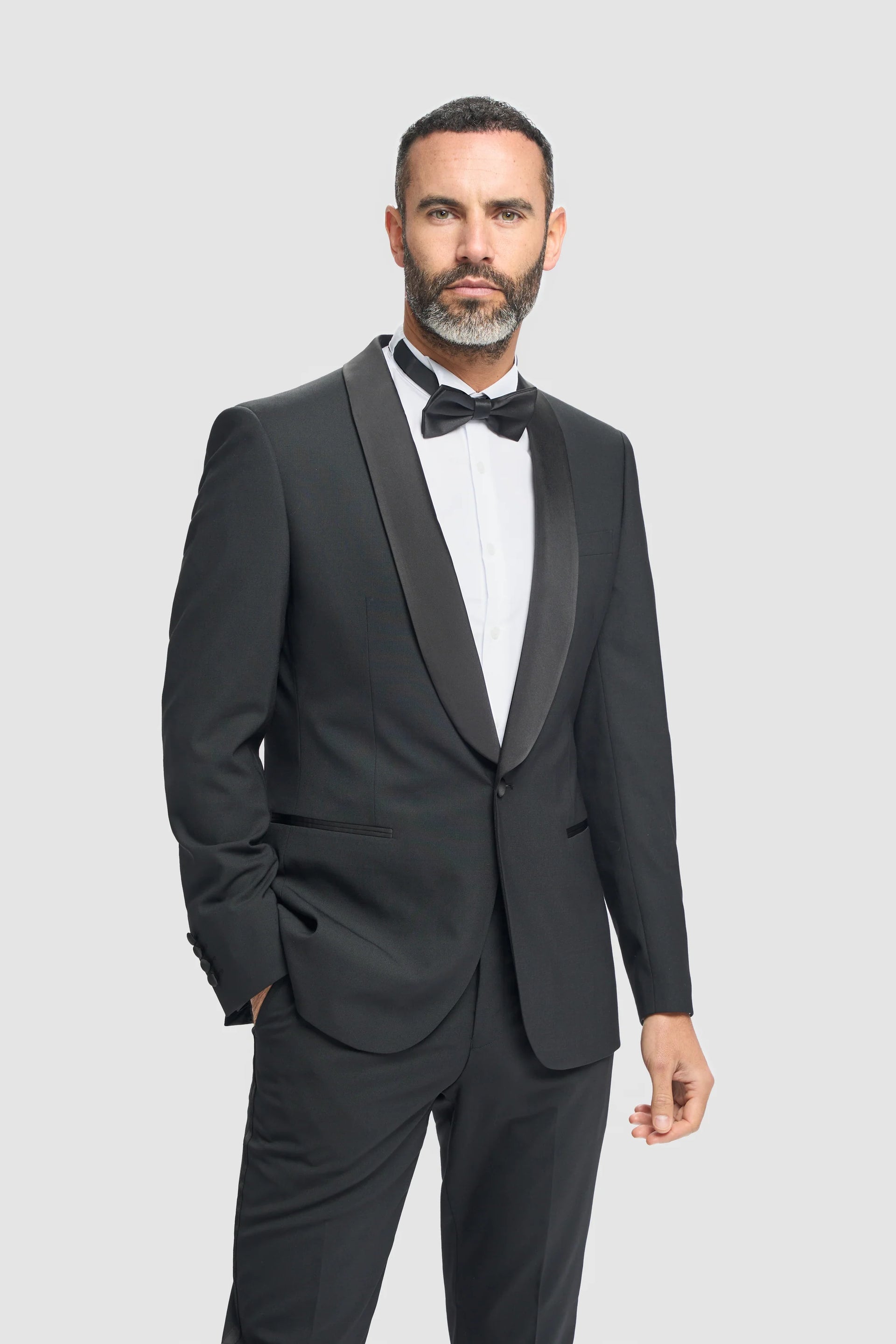 Homem vestindo um smoking preto com camisa branca e gravata borboleta preta sobre um fundo cinza claro.