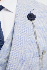 Homem vestindo terno cinza claro com gravata azul-marinho e broche de flor sobre fundo branco.