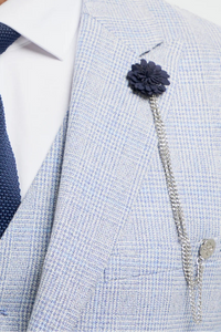 Homem vestindo terno cinza claro com gravata azul-marinho e broche de flor sobre fundo branco.