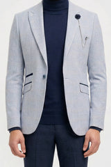 Homem vestindo blazer cinza claro, gola alta azul-marinho e calça escura sobre um fundo branco.