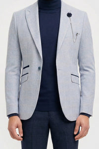Homem vestindo blazer cinza claro, gola alta azul-marinho e calça escura sobre um fundo branco.