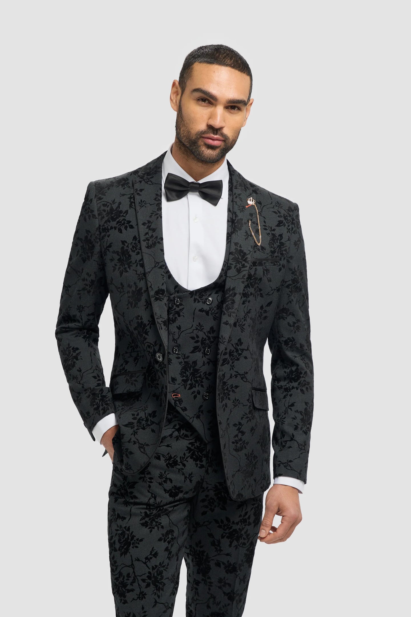 Homem vestindo um terno preto florido com camisa branca e gravata borboleta sobre um fundo cinza claro.