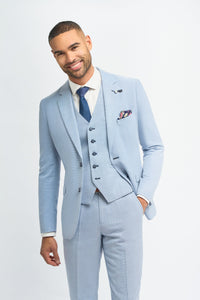 Homem vestindo terno azul claro com camisa branca e gravata azul sobre um fundo liso.